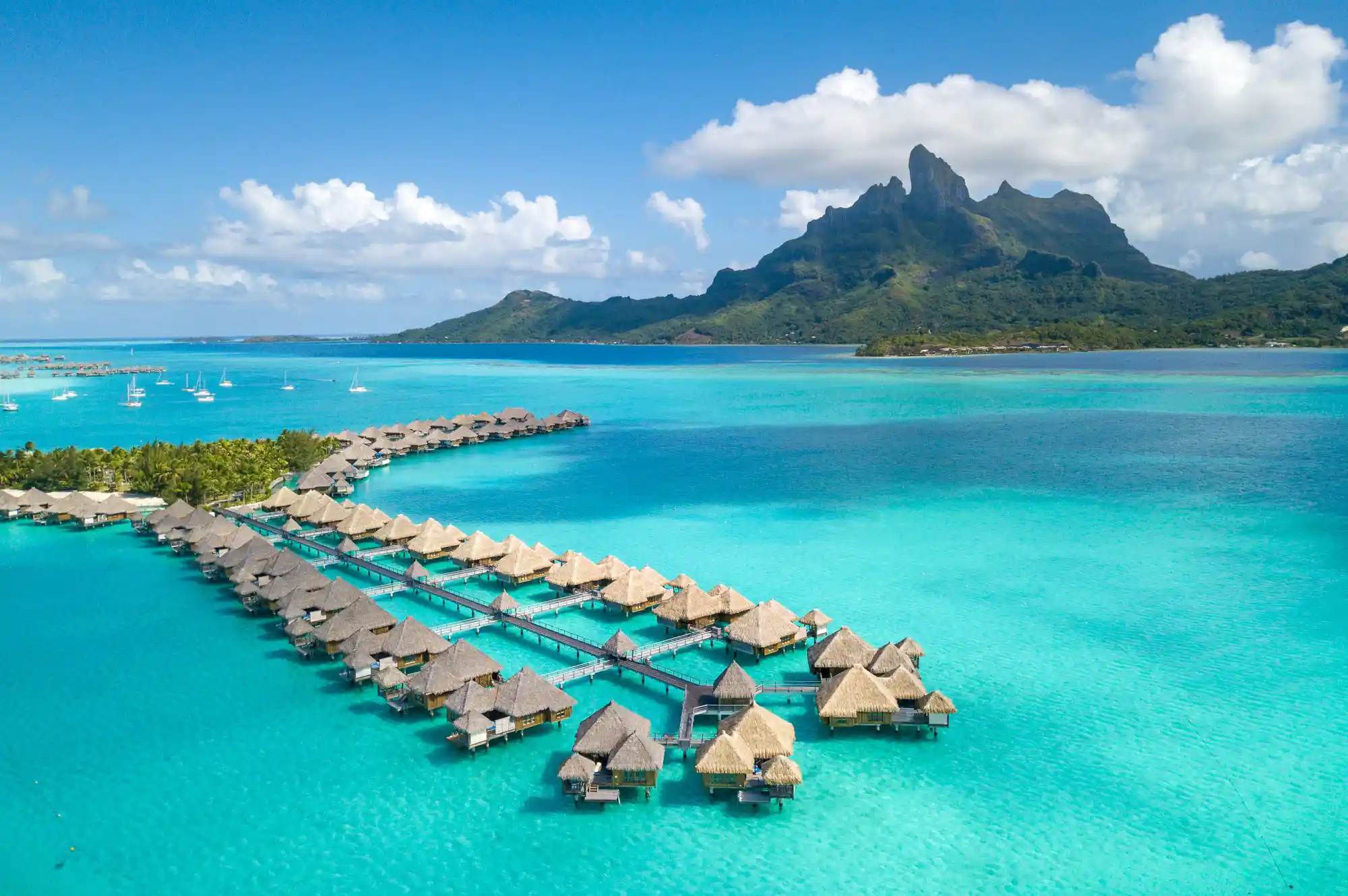 Overwater bungalows situated over a bright blue lagoon with a dramatic mountain peak in the background.