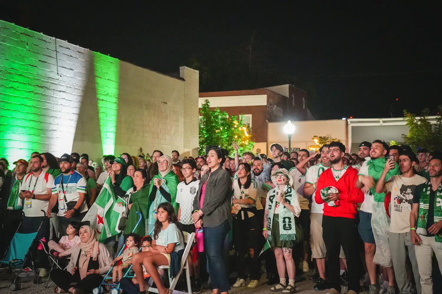 A diverse crowd of soccer fans wearing jerseys and scarves cheering during an Algeria tournament game at an outdoor venue.
