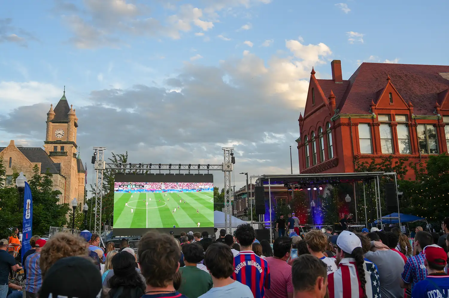 A large crowd gathered in a downtown Lawrence street in front of an outdoor screen showing a soccer match with historic brick buildings behind.