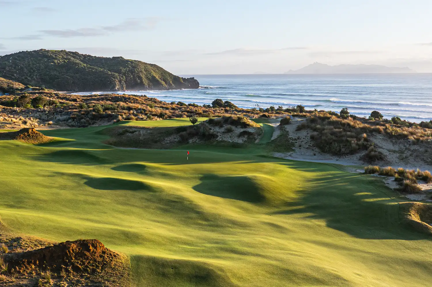 Golf course green with sand dunes and ocean waves in the background.