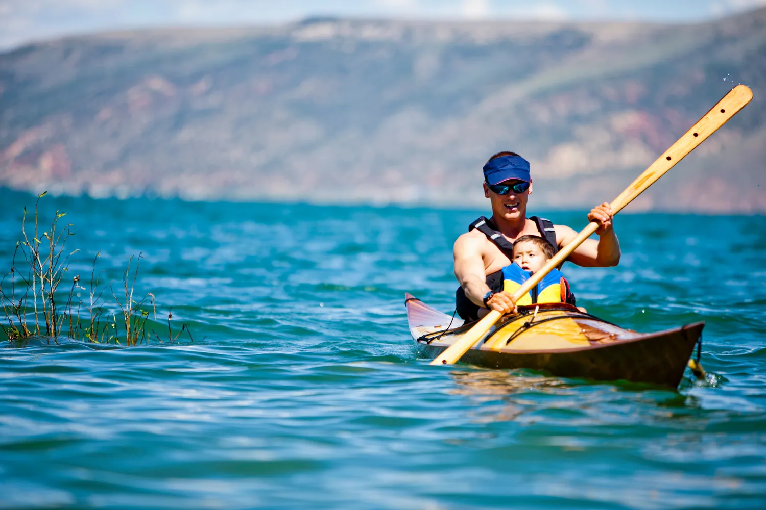 A man and child paddling a yellow kayak on the calm blue waters of Bear Lake with mountains in the background.