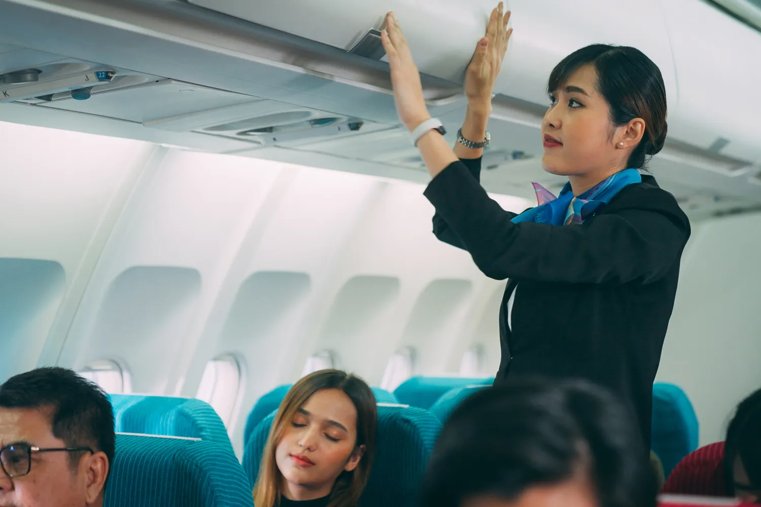 A flight attendant in uniform reaching up to secure an overhead compartment in a crowded airplane cabin.