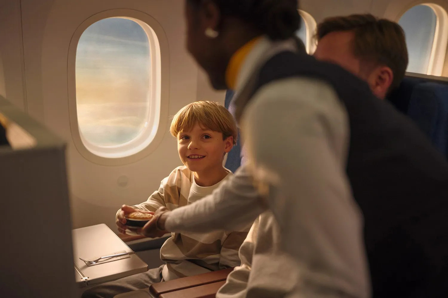 A smiling flight attendant serving a pastry to a young passenger in the aircraft cabin.