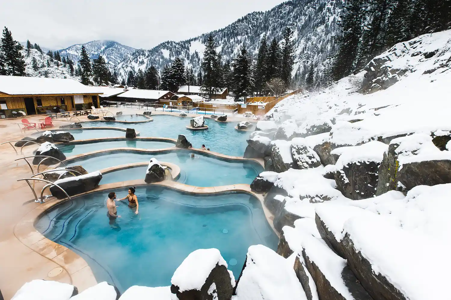 Two people relaxing in outdoor hot spring pools with a backdrop of snow-covered mountains and evergreen trees.