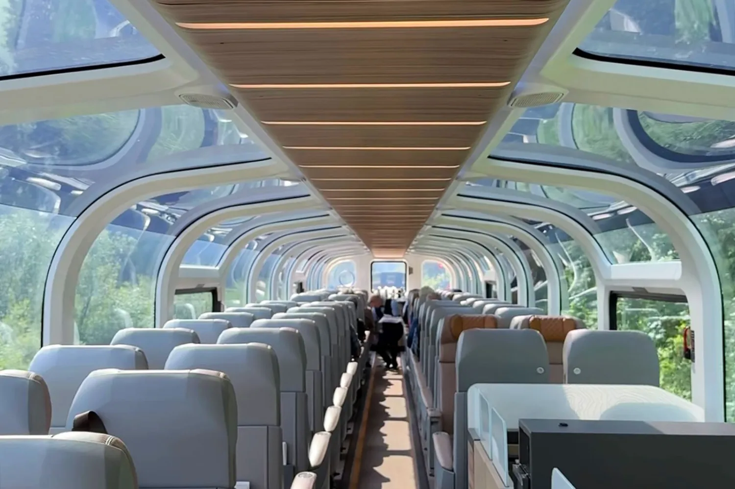 Interior view of a Rocky Mountaineer glass-dome coach with passengers looking out at green forests through panoramic windows.
