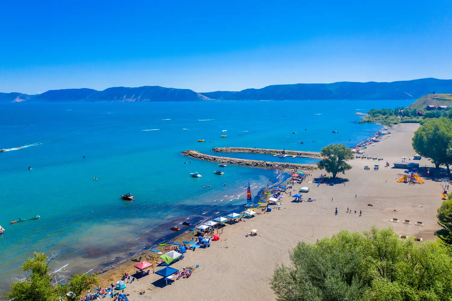 A crowded beach at Bear Lake with people swimming and relaxing near the shore against a backdrop of distant hills.