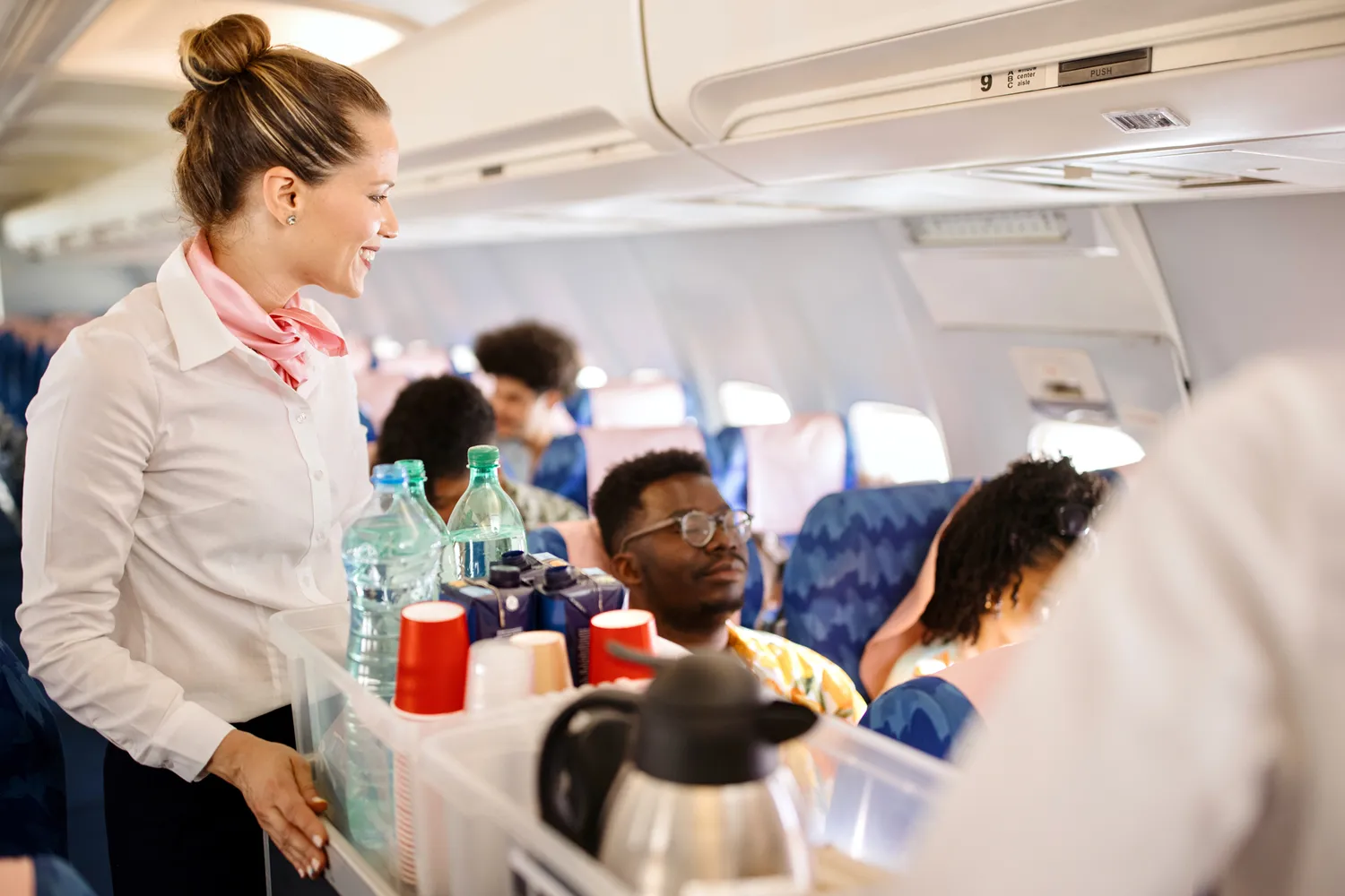 A flight attendant moves a drink cart through the narrow airplane aisle to serve seated passengers.