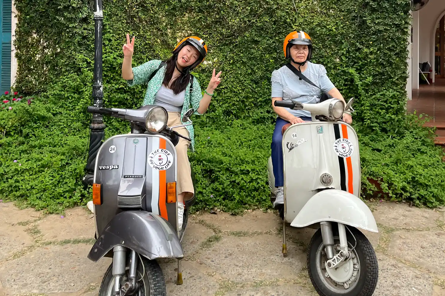 Two travelers on the back of vintage Vespa scooters smiling and gesturing for the camera.