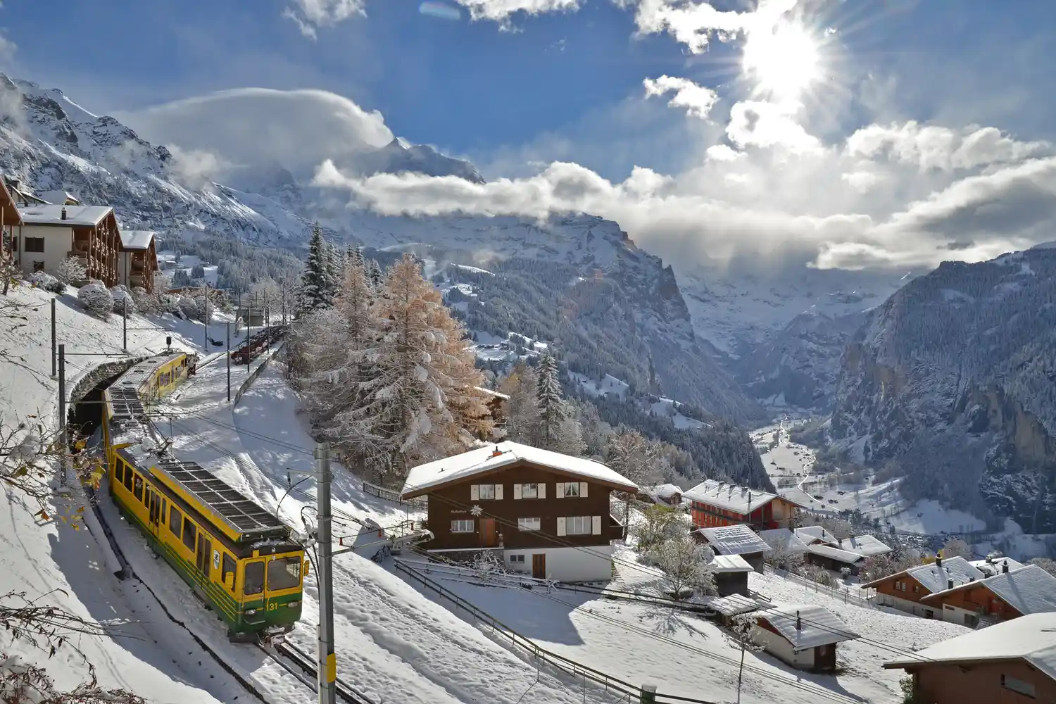 A yellow and red cog railway train winding through a snow-covered alpine village with chalets.