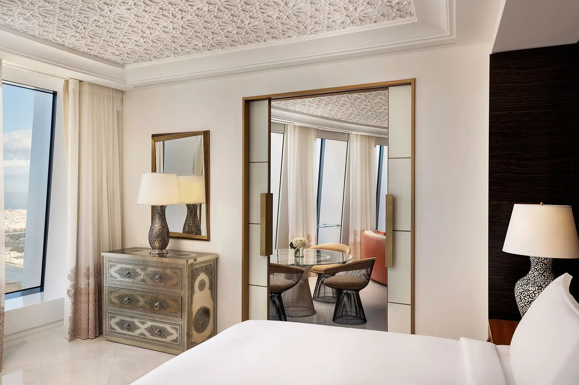A luxury hotel bedroom suite with plush bedding and a soft-lit connecting lounge area.