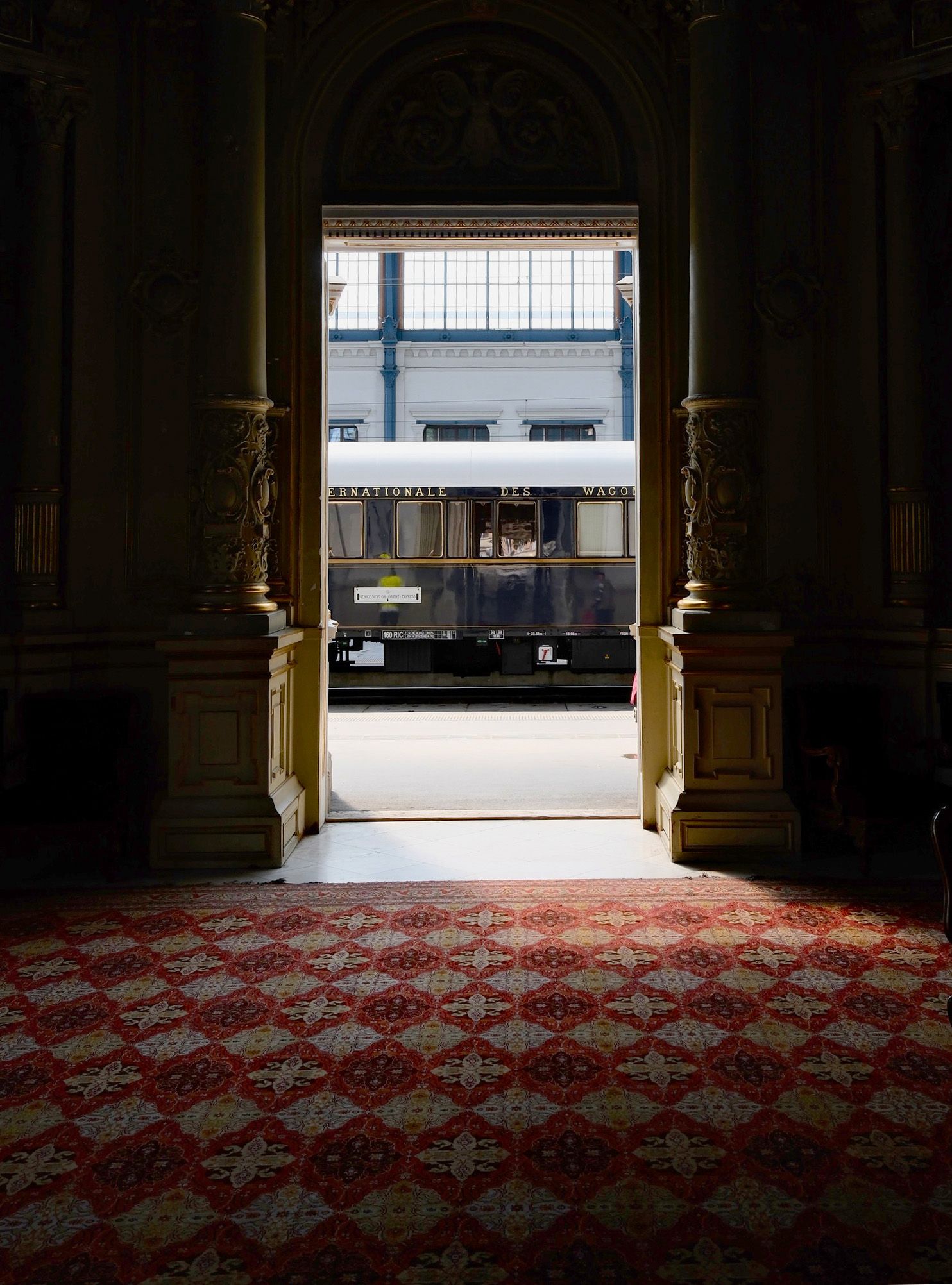 The narrow, polished wood corridor of a vintage train carriage with Art Deco lamps and patterned carpets.